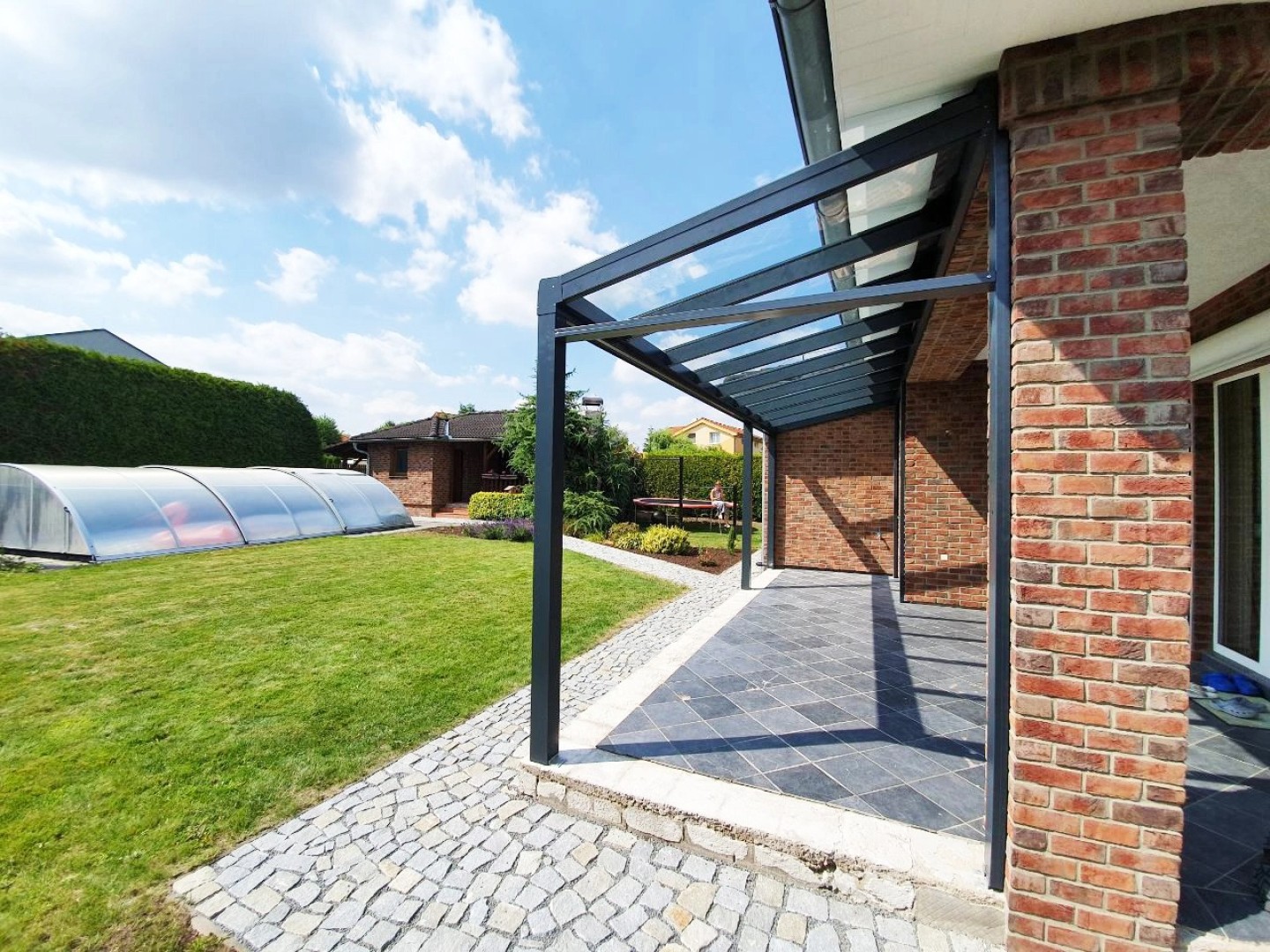 Moderná antracitová hliníková pergola so sklenenou strechou pri tehlovom dome so záhradou a bazénom.